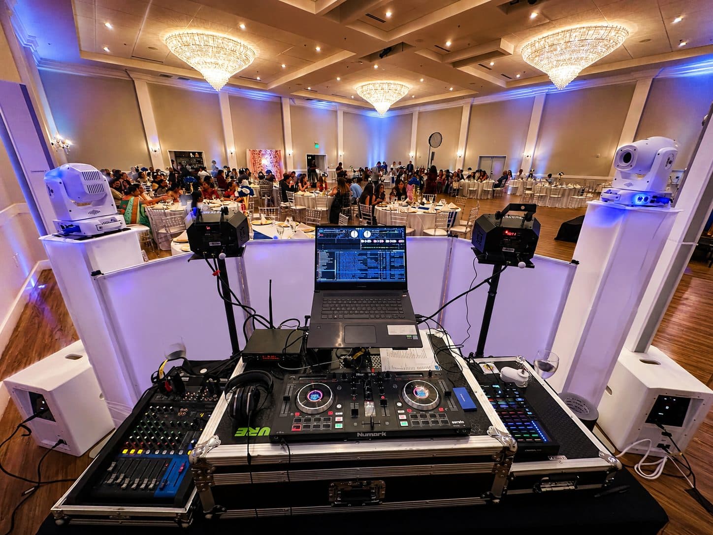 A Vyoma Productions DJ booth with professional decks and moving-head lighting overlooking a chandelier ballroom set for a wedding